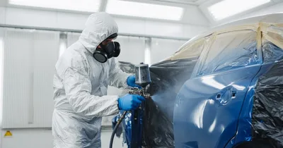Technician in a protective suit and respirator spray-painting a car panel inside a brightly lit paint booth