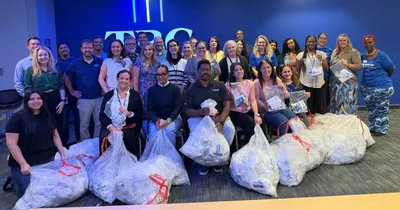 Group of TBC Corporation employees posing with bags of assembled hygiene kits in front of a blue wall with an illuminated TBC sign