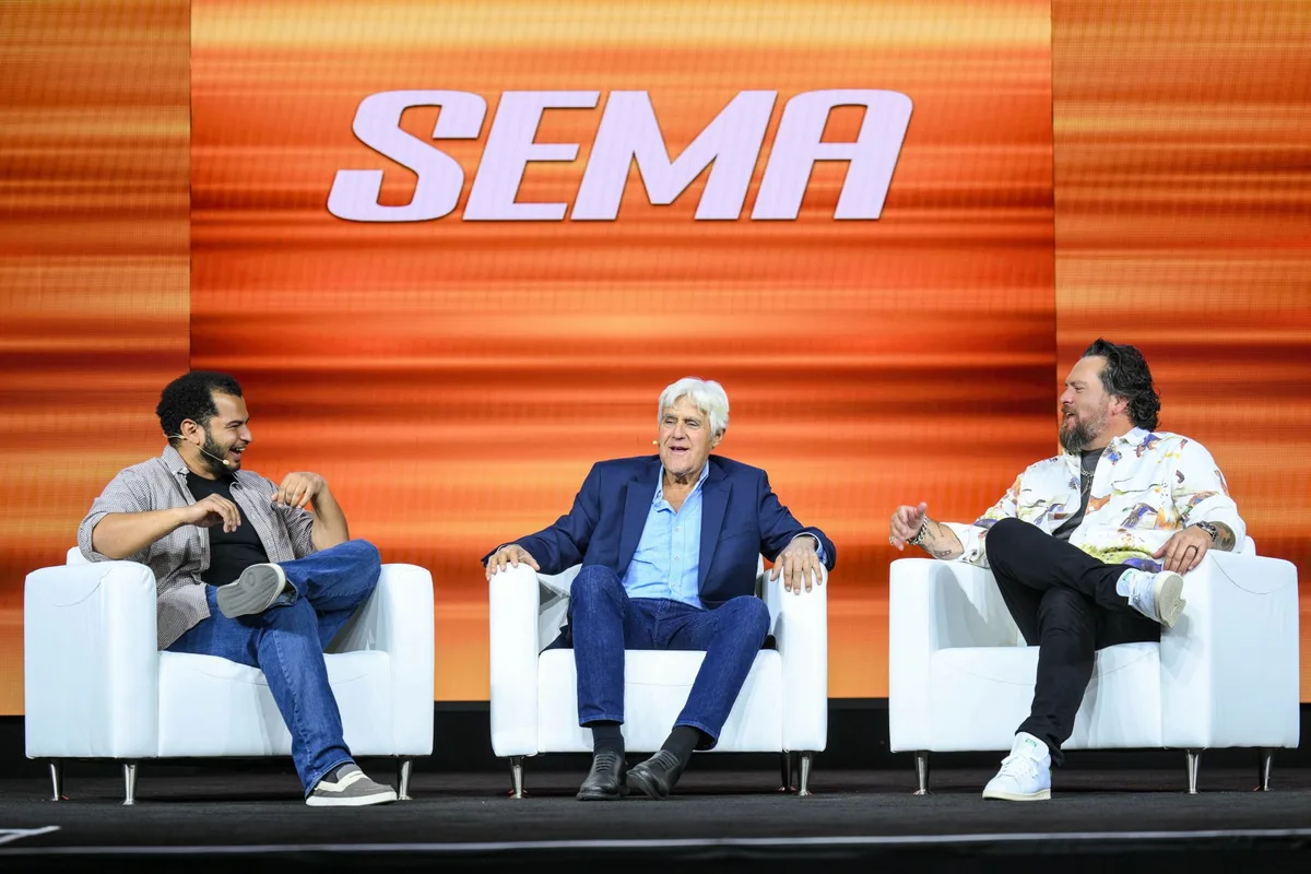 Jay Leno seated on stage in a white armchair between two other speakers, with a large SEMA logo on an orange screen behind them.