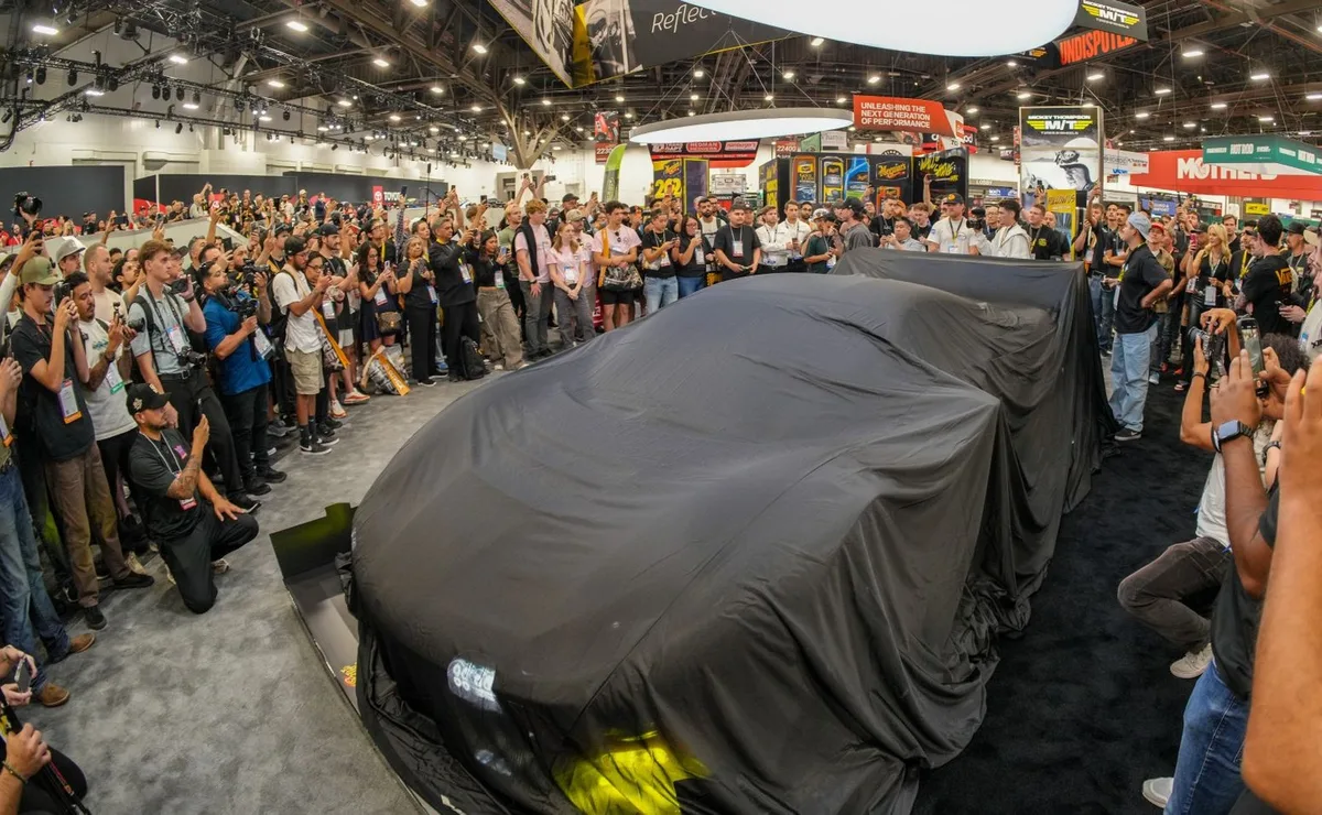Crowd of attendees and photographers with cameras and phones surrounding a vehicle under a black cover before an unveiling on the SEMA Show floor