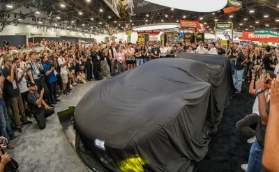 Crowd of attendees and photographers with cameras and phones surrounding a vehicle under a black cover before an unveiling on the SEMA Show floor
