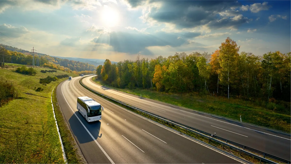 White coach traveling on a curving divided highway through autumn woodland under a sunlit sky