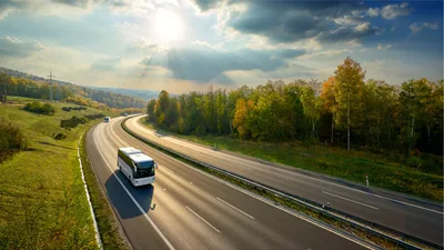 White coach traveling on a curving divided highway through autumn woodland under a sunlit sky