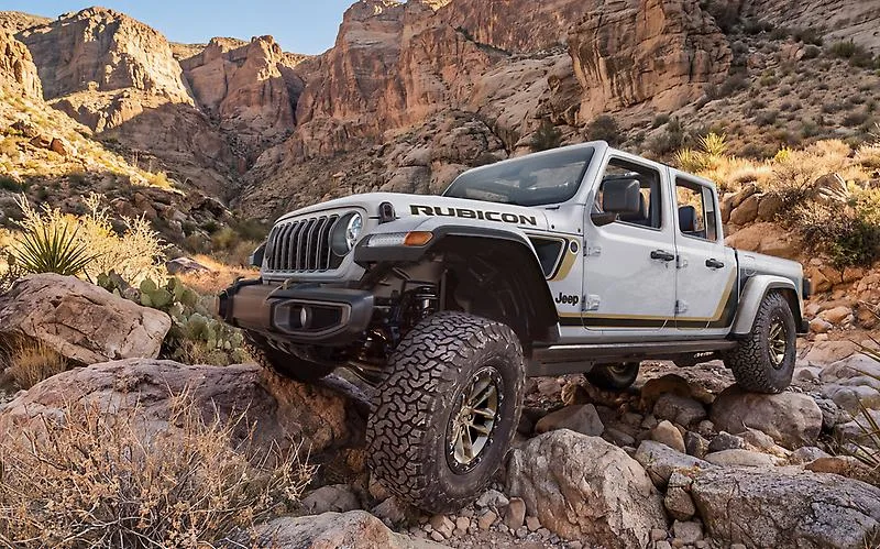 Silver 2027 Jeep Gladiator Rubicon with bronze wheels and 35-inch all-terrain tires climbing a rocky ledge in a desert canyon.