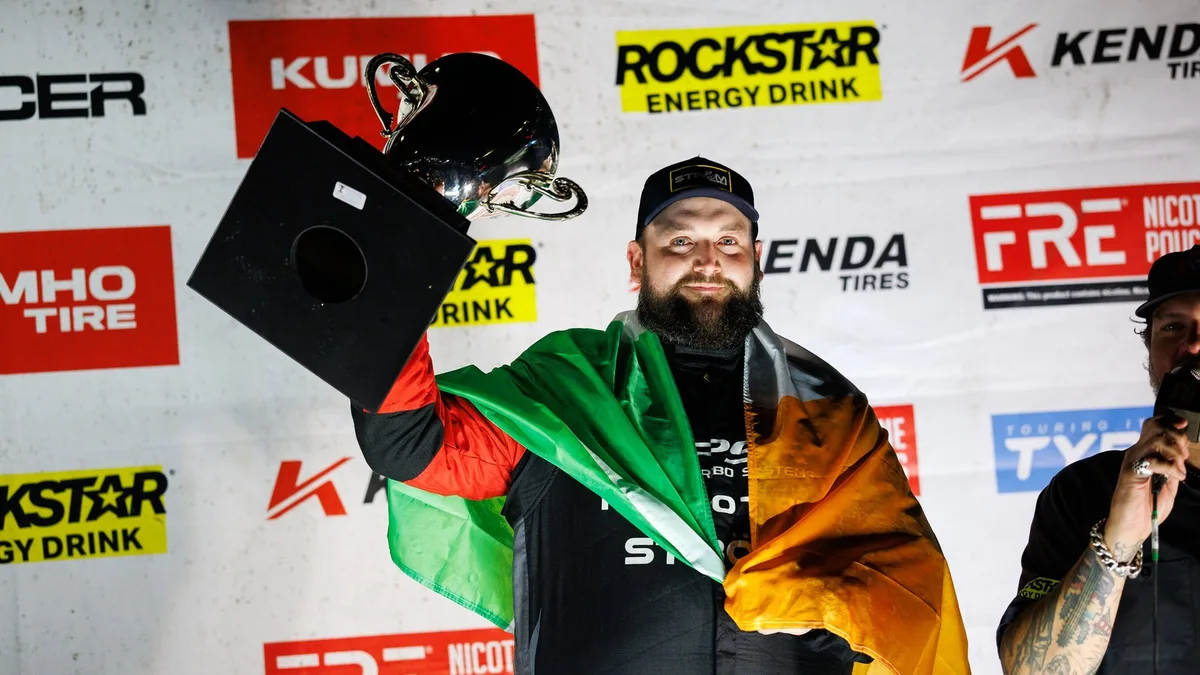 Driver Jack Shanahan, wearing a cap and a racing suit with a green, white and orange flag over his shoulders, holds a trophy above his head in front of a sponsor backdrop.