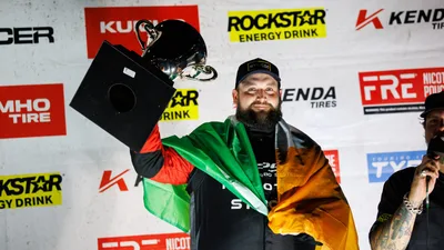 Driver Jack Shanahan, wearing a cap and a racing suit with a green, white and orange flag over his shoulders, holds a trophy above his head in front of a sponsor backdrop.